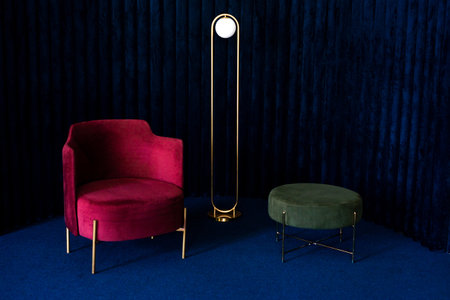 An apartment with a comfortable red velor armchair, a minimalist floor lamp and a green pouffe in a room with dark blue velvet wall panels and floor carpeting. Luxury interior design concept.の写真素材