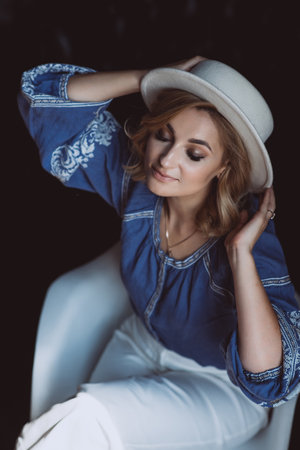 Beautiful stylish elegant blonde woman in ethnic clothes in an embroidered shirt in a white hat in the interior in grunge style. Ukrainian national clothes.の写真素材