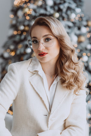 Young happy european businesswoman wearing glasses sitting at workplace with decorated Christmas tree behind, smiling woman working in home office during winter holidays. Soft selective focus.の写真素材