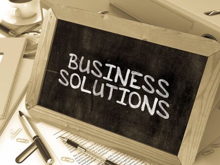 Business Solutions Handwritten by white Chalk on a Blackboard. Composition with Small Chalkboard on Background of Working Table with Office Folders, Stationery, Reports. Blurred Background. Toned Image.の写真素材