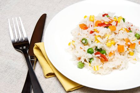 boiled rice with green peas, corn, sweet peppers and carrots on a white plateの写真素材