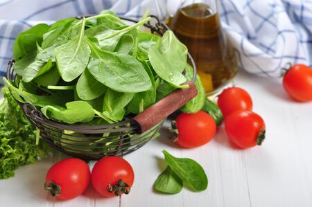 Fresh spinach leaves in basket with tomatoes, cucumber and olive oil on a white wooden backgroundの写真素材