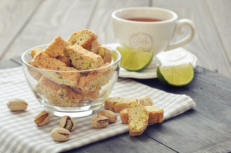Fresh cantuccini with pistachios in glass bowl with cup of tea on wooden backgroundの写真素材