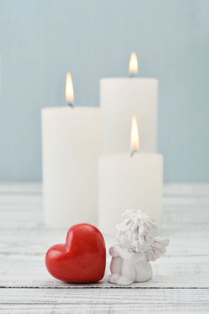 Statuette of small  angel with stone heart and candles over light backgroundの写真素材