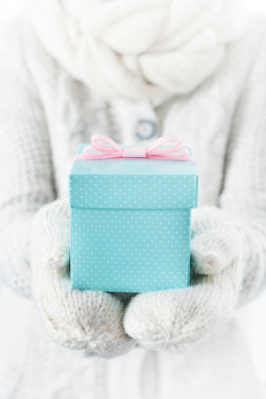 Hands in white mittens holding small blue gift box with pink ribbon.の写真素材