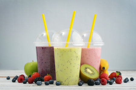 Fruit smoothies in plastic cups with blueberry, strawberry, kiwi, blackberry, raspberry and banana on blue background. Take away drinks concept.