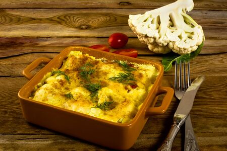 Baked cauliflower in breadcrumbs with cheese on a square ceramic baking dish, fork, knife, tomatoes, wooden background, horizontalの写真素材