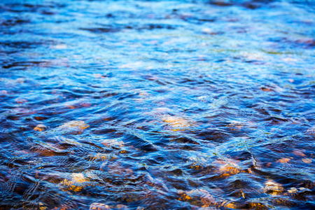Defocused blue water surface background. Stony bottom of a clear mountain river. Flecks of sunlight and ripples on the water of river. Water background.  Water surface texture.の写真素材