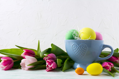 Easter hand painted eggs in blue cup and pink tulips background. Copy space. Happy Easter greeting...