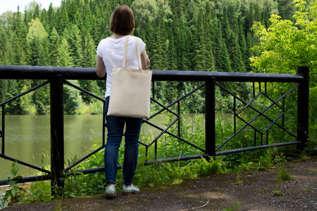 Woman carrying empty reusable shopping bag near bridge railing mockup. Female hand holding canvas eco friendly tote bag mock upの写真素材