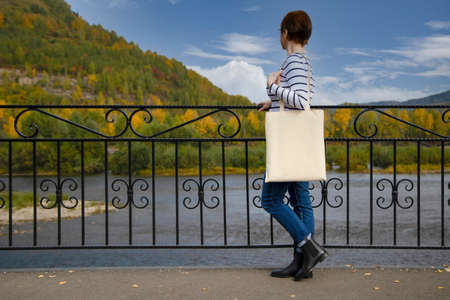 Woman in striped long sleeve shirt carrying empty canvas shopping bag near bridge railing mockup. Female hand holding canvas eco friendly tote bag mock upの写真素材