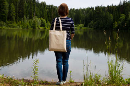 Woman in the striped long sleeve shirt carrying shopping bag mockup, summer river view. Female hand holding canvas eco friendly tote bag mock upの写真素材
