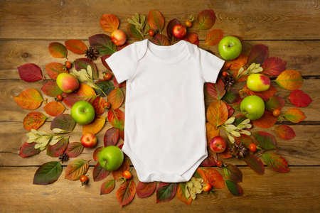 White cotton baby short sleeve apparel rustic mockup with fall leaves, apples and pine cones. Design...