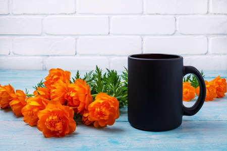 Black coffee mug mockup with orange globeflowers on the blue table. Empty mug mock up for brand...