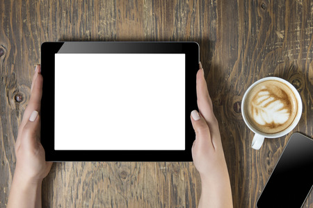 Businesswoman reading his tablet with white screen and a cup of coffee smartphone in coffee cafe. Woman hands holding tablet pc with empty screen on wooden backgroundの写真素材
