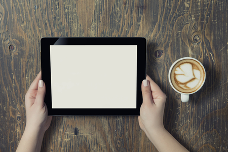 Businesswoman reading his tablet with white screen and a cup of coffee smartphone in coffee cafe. Woman hands holding tablet pc with empty screen on wooden backgroundの写真素材