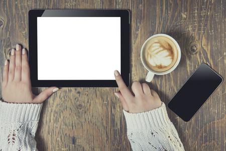 Businesswoman reading his tablet with white screen and a cup of coffee smartphone in coffee cafe. Woman hands holding tablet pc with empty screen on wooden backgroundの写真素材