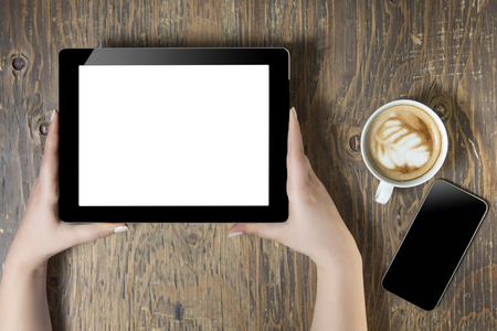 Businesswoman reading his tablet with white screen and a cup of coffee smartphone in coffee cafe. Woman hands holding tablet pc with empty screen on wooden backgroundの写真素材