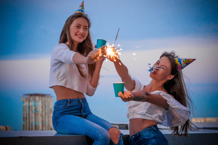 Two girls with Bengal lights and paper glasses celebrate a birthday against the sky