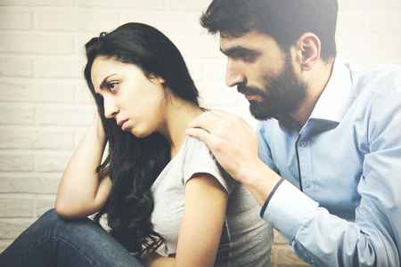 Depressed young woman looking away while man sitting behind her on the couch and consoling herの写真素材