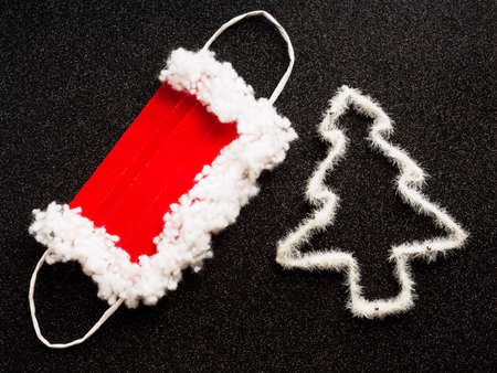 Flat lay of protective mask for Santa Claus (red and white) and one white Christmas tree on black...
