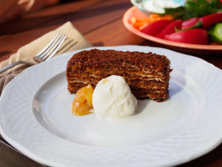 piece of cake brownie on a white plate with honey and a scoop of ice cream on the background of a plate with vegetablesの写真素材