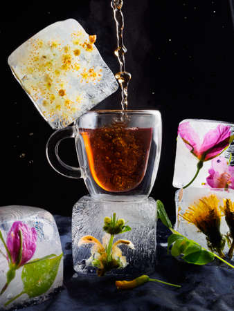 a transparent cup of tea in ice, ice in the form of frozen cubes with flowers, hot tea is pouring,...