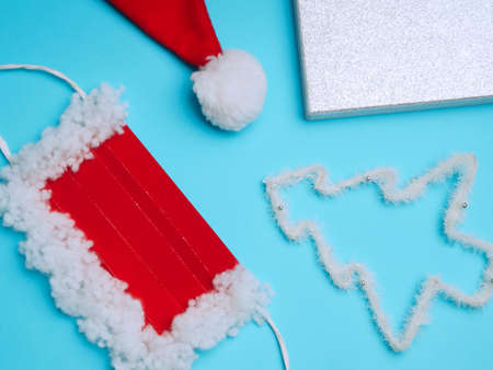 Flat lay of protective mask, part of a hat with a pompom for Santa Claus, Christmas tree, gift wrap. Red and white on a blue backgroundの写真素材