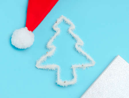 christmas concept - flat lay of Santa Claus hat pompom, silhouette of a white Christmas tree made of fluffy wire and silver gift box on a blue backgroundの写真素材