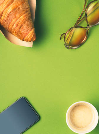 Flat lay paper cup of coffee, croissant, phone, sun glasses. Breakfast concept. Croissant is wrapped...