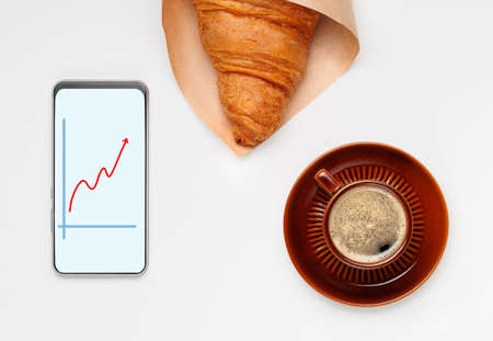 Flat lay of brown cup of black coffee with saucer, croissant and phone. Croissant wrapped in craft paper, abstract stock market chart in red on phone screen. Investor breakfast concept. Minimalism.の写真素材
