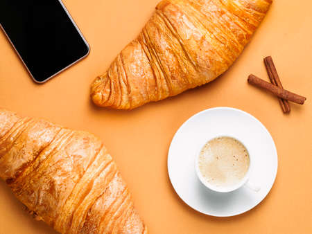 Flat lay of white cup of coffee, two croissants, cinnamon sticks, phone. Brown paper background. Breakfast concept. Cup and saucer.の写真素材