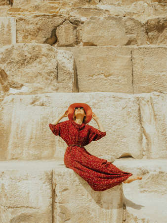 A Caucasian woman in a red dress and hat sits beautifully on the lower blocks of the Khafre pyramid...