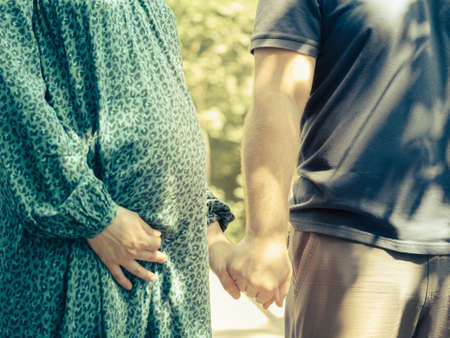 Man and woman hold hands in nature on a sunny day. The woman is pregnant. A pregnant woman is dressed in a green dress, supporting her belly with one hand. Pregnancy and motherhood concept. Close-up.の写真素材