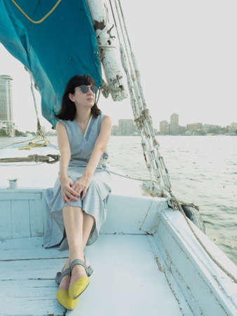 Portrait of a woman on a felucca. A Caucasian woman in a blue dress and sunglasses sits beautifully in a felucca boat in the evening sun against the backdrop of Cairo in Egypt on the Nile River.の写真素材