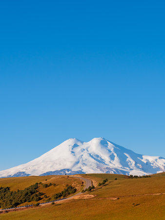 A beautiful asphalt road through the autumn hills of the Caucasus leads to Elbrus. Elbrus with two peaks against the blue sky is covered with snow. Copy space.の写真素材