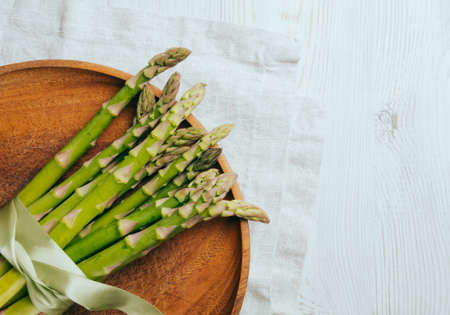 A bouquet of green fresh asparagus is tied with a green silk ribbon. Asparagus lies on a round wooden plate on a light table with a linen napkin, top view. Copy space.の写真素材