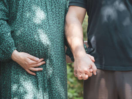 Man and woman hold hands in nature on a sunny day. The woman is pregnant. A pregnant woman is dressed in a green dress, supporting her belly with one hand. Pregnancy and motherhood concept. Close-up.の写真素材