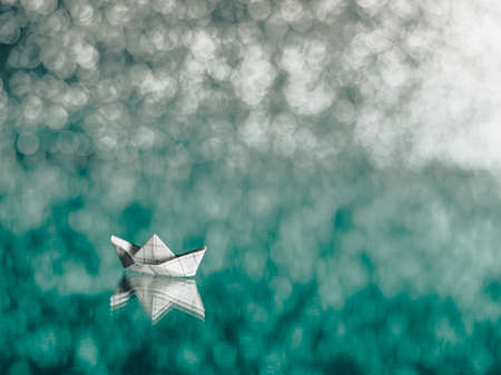 Paper boat from a math notebook. The origami ship floats on a mirrored surface like on the blue sea, all in beautiful defocused silver lights and bokeh. Copy space.の写真素材