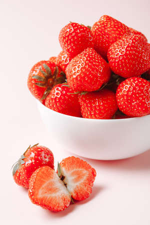 whole ripe strawberries in a white bowl on a pink background, a couple of berries lying side by...