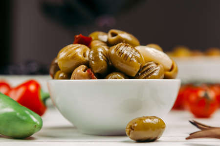 grilled green olives in oil, pitted, with red hot pepper and garlic in a white bowl, still life with tomatoes, glasses of red wine, wooden skewers for olives, side viewの写真素材