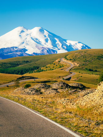 A beautiful bend of the road through the green hills of the Caucasus to the main mountain - Elbrus. Elbrus with two peaks against the blue sky is covered with snow. Copy space.の写真素材