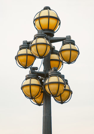A vintage streetlight pole with multiple round caged lights illuminates a cloudy day. The pole is...