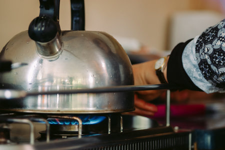 A stainless steel kettle with whistle on a gas stove and the hand of a woman who has just turned on...