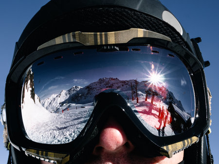 Black helmet and ski goggles on a man. The blue sky, snow hills, slopes, ski lift and people reflect...