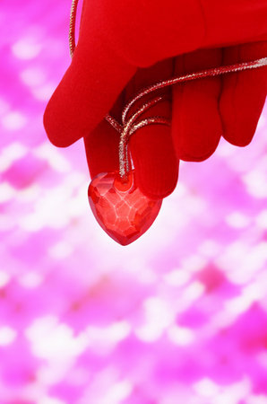 Hand in a red glove holding a heart with a golden rope Abstract pink background with bokeh effect and glitter. St. Valentine's Day background.の写真素材