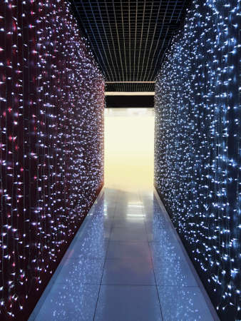 A dark, long corridor illuminated by many light bulbs with blue and pink glow, and at the end of the...