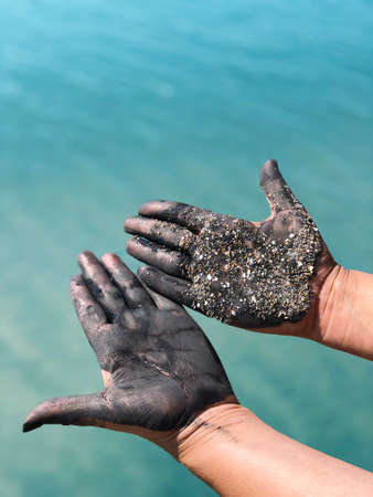 Hands with palms up, stained with black mud and sand, against a background of turquoise water.の写真素材