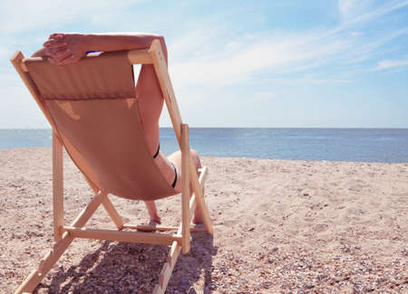 Young woman on the beach, sitting back in a chair, hands up and looking at the sea. Summer beach...