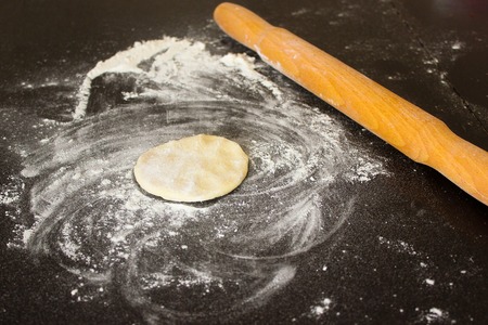 Small round dough piece with flour and rolling pin on black kitchen table. Shot of prepairing of...